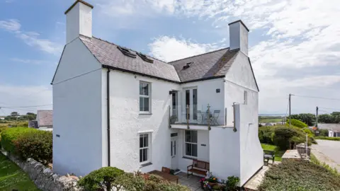 Michael Speak The outside of a two-story L-shaped white-wash house. There is a small balcony at the front and a wooden bench and plantpots outside. It is surrounded by grassy gardens and farm buildings. 