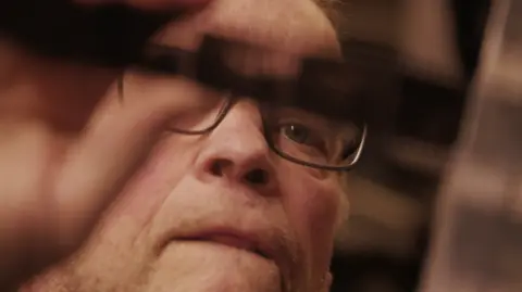 Firecrest Films Close up of a bald man wearing glasses looking intensely at a camera film negative.