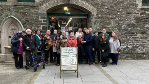 BBC A group of traders gather outside of Tiverton Pannier Market. The group, made up of about 20 people, are standing in front of a stone wall. A sign which shows the market's opening hours is in front of the group. 