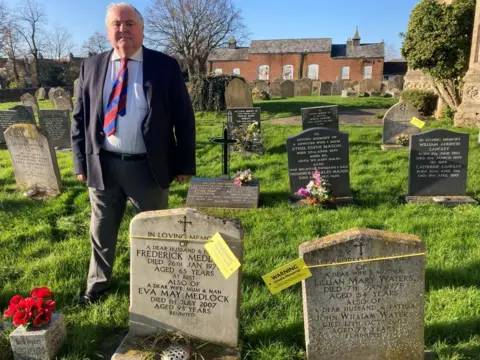 Jill Bennett/BBC Councillor Paul Kunes stood behind graves with yellow tags