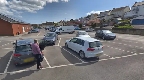 Google A man leans on his car in a car park full of cars on a sunny day