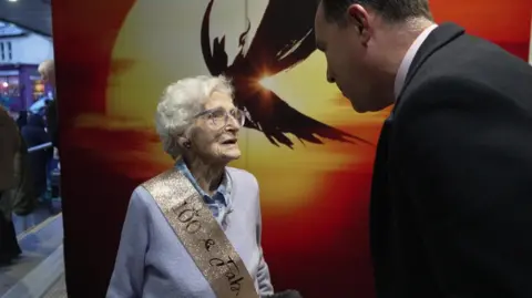 Cilla talks to Ed. A Miss Saigon poster is in the background.