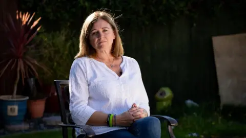 PA Media Debbie Adlam, a woman with short, light brown hair, sits on a garden chair. She is wearing a white top with three-quarter length sleeves and a pair of blue jeans. She is pulling a neutral expression and her hands are clasped together in her lap.