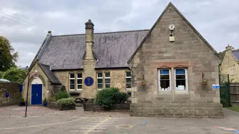 The picture shows Eyam Church of England Primary School. It's an old style school building with a blue door to the left and and the playground at the front with a netball post.