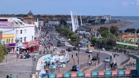 Marine Parade in Southend-on-Sea on a sunny day with a clear blue sky. People are walking along the pavement and cars being driven along the road. There are amusements to the left and a theme park to the right.