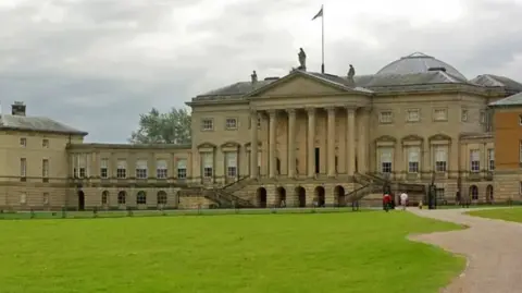 The imposing columned frontage of an 18th Century stately home, with a grand entrance on the first floor led to by stairs, and a manicured lawn in front