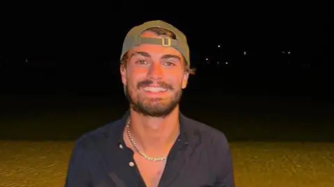 Rockdale Ilinden FC Dan Elkayam smiling against a black background. He is a young man with a short brown beard and moustache, and wears a green sunhat backwards on his head. He is wearing a blue buttoned shirt and a shell necklace.