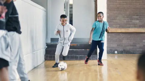 The Devas Club Two boys playing football inside, one wearing a pale tracksuit and smiling as he's running with the ball in front and another child wearing a blue tshirt with the words awesome on it is alongside, smiling. 