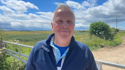 A man with grey hair, dressed in a blue zip-up jacket over a white and blue top, stands by a metal farm gate along a dirt track, with fields stretching into the distance and power lines visible under a bright, cloudy sky.