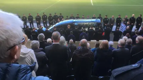 BBC People standing behind a funeral cortege, driven pitch side