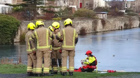 Wolf Gruellich Six fire fighters in a huddle on the banks of the pond. They are wearing yellow uniforms and helmets.