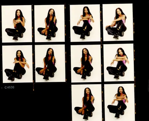 RANKIN Contact sheet of multiple film photographs of a young girl with long, braided hair. Wearing a pink top and black trousers, posing in a squatted, cross legged position
"Samantha Mumba, Press Images, 2000, (c)Rankin"