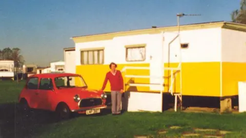 Richard Dowell An old photo of Richard, wearing a red jumper and brown trousers, standing with a red mini on his left and a brightly painted static caravan on his right, The van is mustard yellow on the bottom and white on the top, with a mustard trim on the roofline