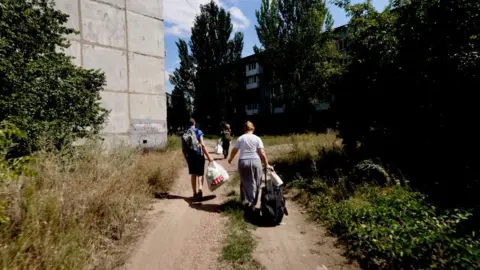 Trīs cilvēki staigā pa netīrumu ceļu garām ēkai un nezāļu kaudzēm, pārvadājot lielas somas