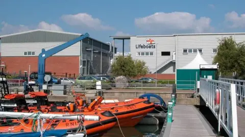 Lukas Antczak A picture taken on a jetty shows three orange RNLI lifeboats docked side by side. Beyond the boats, a large white building bears a sign with the word "Lifeboats" underneath a St George's Cross flag. Several cars can be seen parked in a car park at the front of the building. 