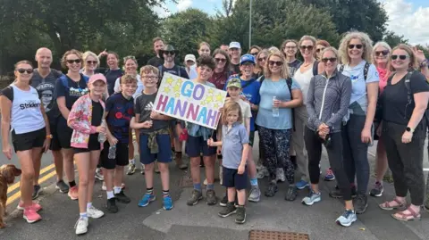 About 30 people stand around in running gear. In the middle a child holds a homemade banner that says Go Hannah