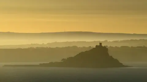 St Michael's Mount viewed across the bay during a yellow sunrise. The land behind it is swathed in mist and the castle is in silhouette.