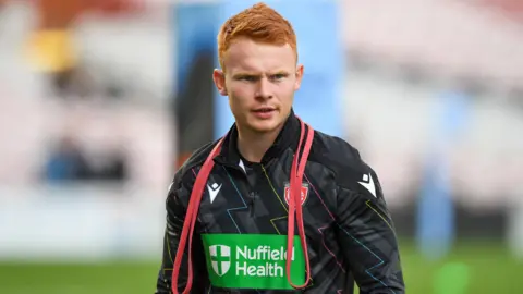 Scrum-half Caolan Englefield warming up for a Premiership Rugby match with Gloucester, wearing a black training top, with red stretch bands around his neck. 