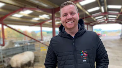 A smiling man in a Dalscone Farm jacket stands in a large agricultural shed