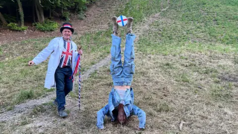 A young woman wearing blue denim jeans, a blue denim jacket, and brown boots does a headstand as she balances a wheel of Double Gloucester cheese between her feet. A man wearing a Union Jack top, a white coat, and a black top hat with red, white and blue ribbons around it stands beside her.