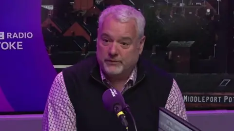 A man with white hair, wearing a checked shirt and a gilet, speaks into a purple microphone. He has a purple background behind him.