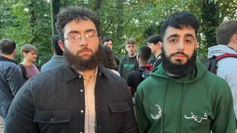 Qays Najm/BBC Noordin Jamal on the left and Sharief Younis stood to his right. Both men are looking at the camera with solemn expressions and behind them is a crowd of other students engaged in a conversation. Noordin Jamal wears a navy jacket with a beige shirt underneath and has thin rectangle framed glasses. Sharief Younis is wearing a dark green hoodie.