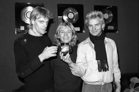 Getty Images [L-R] Stewart Copeland in black top, Andy Summers (in black top) and Sting (in white top and dark scarf) -- holding a glass and smiling, at the A&M offices after signing their record deal in 1978