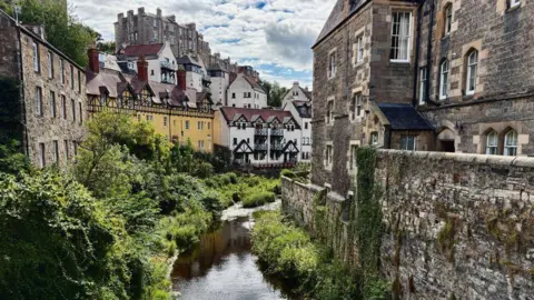 Dwight McAnear A narrow river winds through a lush green corridor, bordered by stone buildings and traditional timber-framed houses. The scene features historic architecture, ivy-covered walls, and a partly cloudy sky.