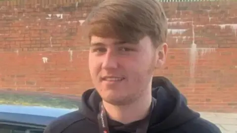 Handout Ellis Cox smiles at the camera wearing a black hoodie in front of a car on a road in front of a red brick wall