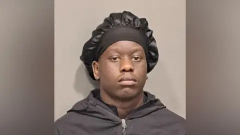 A head-and-shoulders portrait shows a person facing the camera against a plain grey background. The person is wearing a dark zip-up hoodie and a black satin bonnet with a wide headband. The image is evenly lit, with the subject positioned centrally in the frame. No text, logos, or other objects are visible in the photograph.