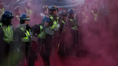 Several police officers in riot gear, clouded by red smoke from a flare
