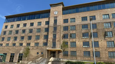 Large brown building with central column with a clock face at the top. Steps at the bottom leading to double door.