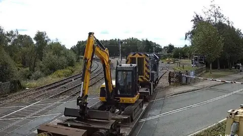 RAIB A yellow digger loaded onto a flat, open railway car. It's arm is bent with the bucket resting on a raised platform on the car. A yellow and black engine is visible. The picture is taken at a level crossing and railway tracks, an intersecting road and greenery are visible. 