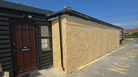 The Pilot Inn A single storey building covered with sheets of chipboard. Guttering runs down the side of the chipboard, which is next to a brown door with a sign reading 'Entrance'.