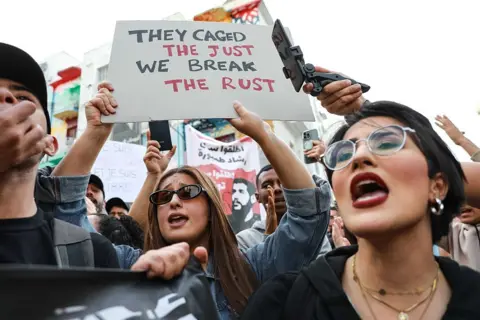 CHEDLY BEN IBRAHIM / GETTY IMAGES A female demonstrator raises a placard reading ''the caged the just, we break the rust'' while demonstrators shout slogans against Tunisia's President Kaïs Saïed.