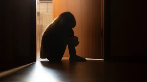 Despairing anonymous child sitting with head on knees in the dark frame of a doorway, backlit by a room behind flooded with daylight 