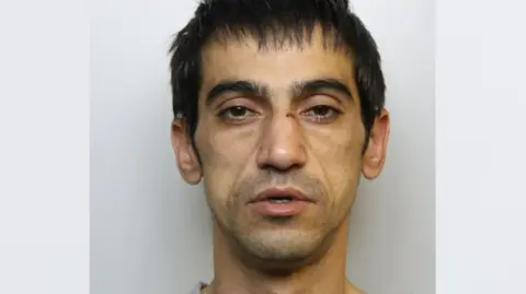 A mugshot of a thin man wearing a grey jumper. He has dark hair, stubble and a cut on his nose.
