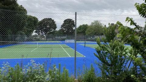 GUY CAMPBELL/BBC Green tennis courts on a blue foundation are framed by verdant undergrowth