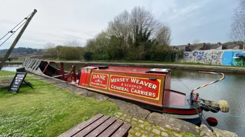 Middleport Pottery A red narrowboat with writing on it reading "Dane" and "Mersey Weaver and S.C.C.Co Ltd General Carriers" in a canal. A picnic table, a black sign and grass are on one side of the canal while a wall and trees are on the other side.