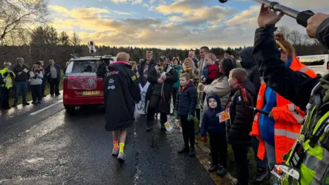 A woman facing away from the camera wears a big black jacket, colourful socks and trainers. Adults and kids stand around on either side of the road smiling at her. A number of them wear headbands will yellow ears on them. 