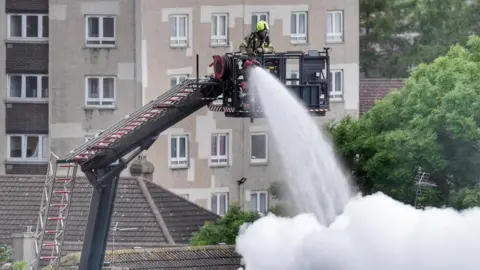Dennis Conaghan Firefighter on turntable ladder putting water on fire from above. There is lots of smoke. 
