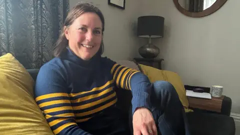 A woman with dark brown shoulder-length hair sits in a living room on a sofa with green-yellow cushions on it. She is wearing blue jeans and a dark blue sweater with horizontal yellow striped across the body and arms. She is smiling at the camera.
