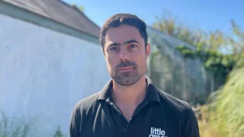 A man with short black hair and stubble looks at the camera, wearing a black polo top with "little green" branding on it.