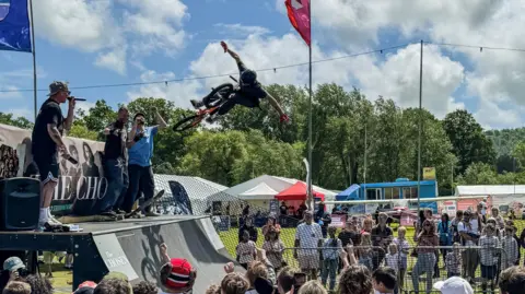 Sunderland BID A BMXer performing a no hander in the air off a quarter pipe at an event. A crowd of people watch while announcers with microphones stand on top of the quarter pipe.