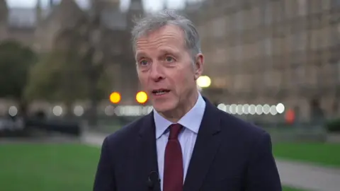 Matt Rodda is standing in front of the Houses of Parliament, wearing a dark suit and a marron tie.