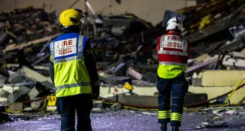 Government of Jersey Two firefighters looking away from camera with pile of debris in the background.