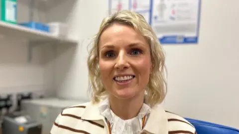 BBC/Helen Richardson Dr Gilmore has blonde, shoulder length curly hair. She is wearing a brown and white striped cardigan and white blouse. She is sitting in her consulting room.