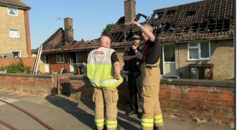 Two firefighters survey the scene with fire-damaged bungalows in the background