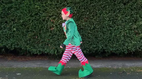 William strides along the pavement in his elf costume, which is green with red and white stripy trousers. He also wears a red and green hat, with large elf ears attached. 