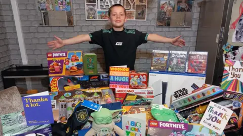 Family photo A wide shot of Phoenix standing in front of a range of presents, including a PlayStation 5, Lego, cards and a Manchester United shirt.
